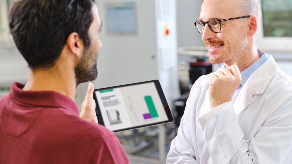 Two people, a nephrologist and a technician, in a discussion in a water treament room. The nephrologist is wearing a white lab coat and the other holding a tablet displaying resource consumotion in dialysis.